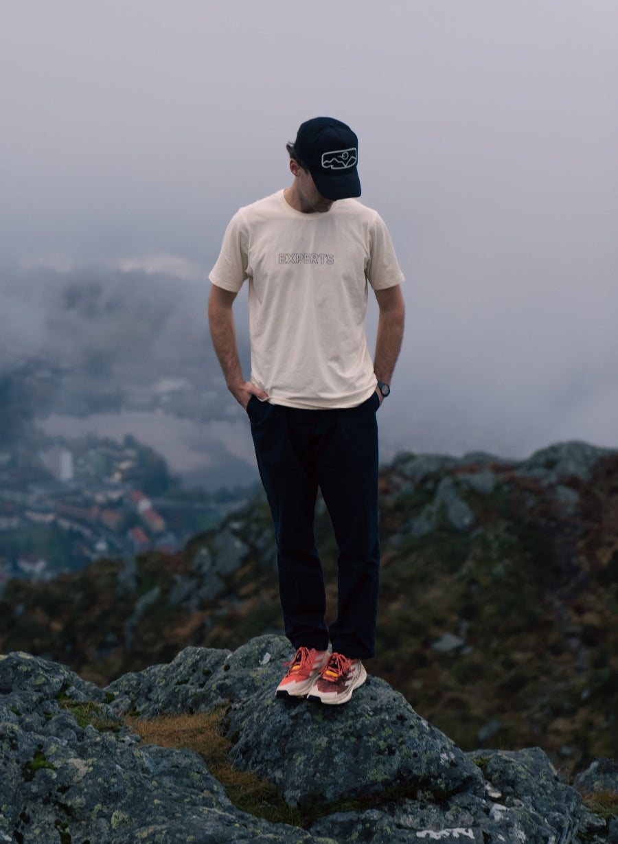 A man stands on a rocky mountain peak wearing a beige t-shirt with 'COAST' printed on the chest, black sweatpants, a black cap, and red sneakers, with a misty mountain valley in the background.