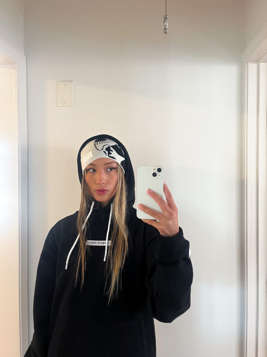 A woman taking a mirror selfie wearing a black pullover hoodie with white drawstrings and a white graphic beanie. The hoodie features a white horizontal text band across the chest.