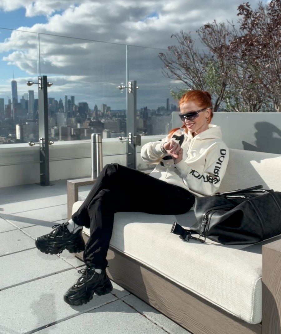 A woman with red hair and sunglasses sits on a modern outdoor sofa on a rooftop terrace, wearing a white hoodie with black text on the sleeve, black pants, and chunky black sneakers. A black bag rests on the sofa next to her, with a city skyline and cloudy sky in the background.