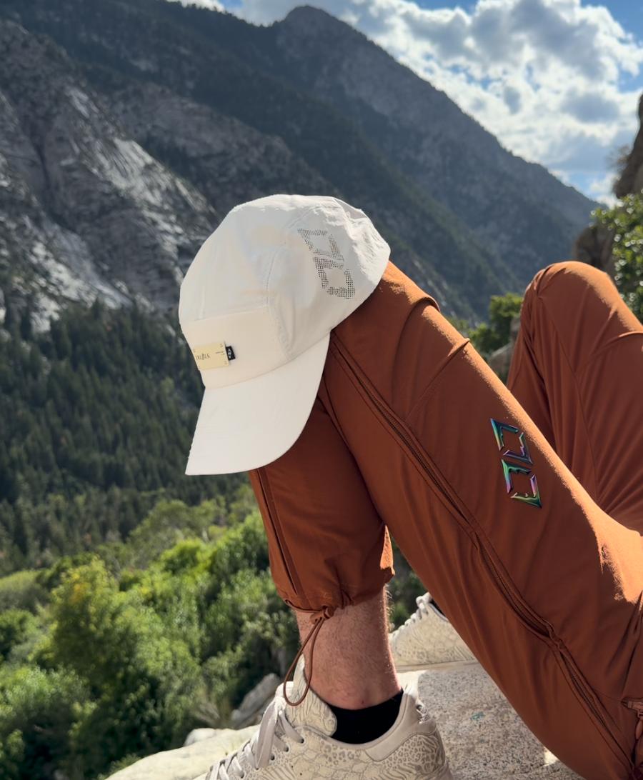 A person outdoors in a mountain landscape, wearing a white baseball cap, rust-brown pants, and off-white sneakers.