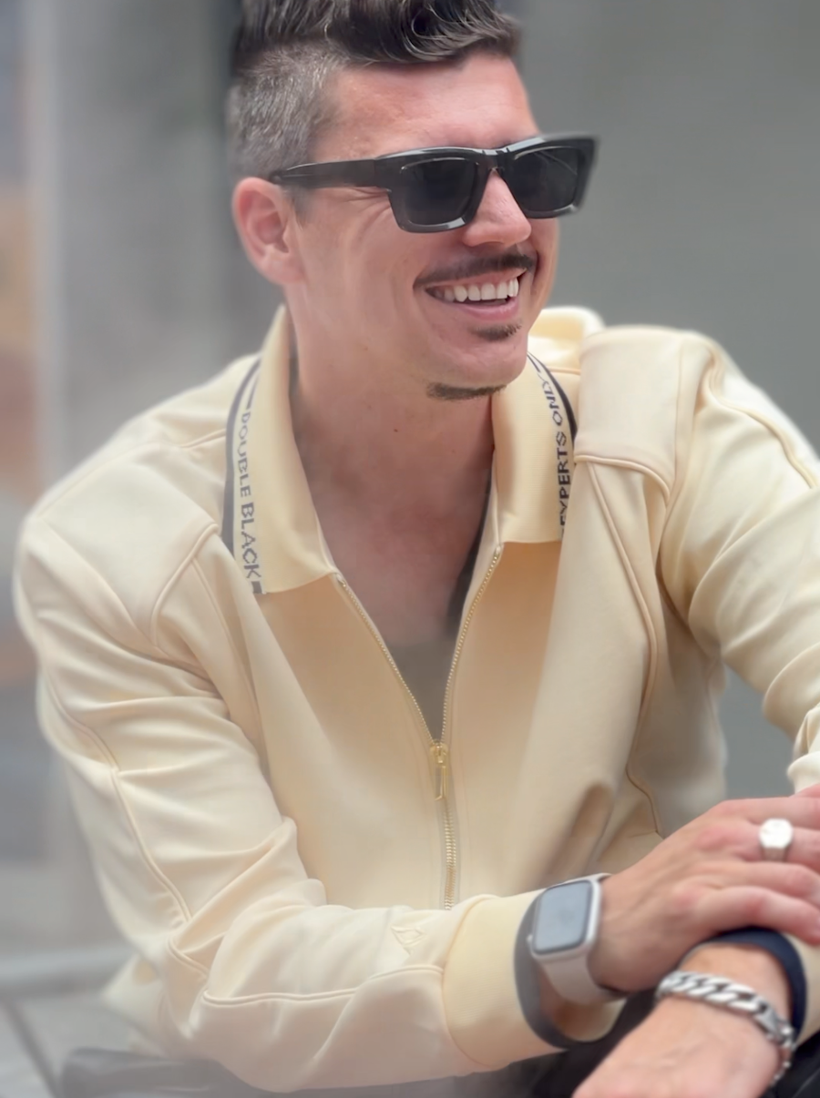 A smiling man wearing black sunglasses, a cream zip-up jacket, and a silver watch, photographed in a real-world setting with a blurred background.