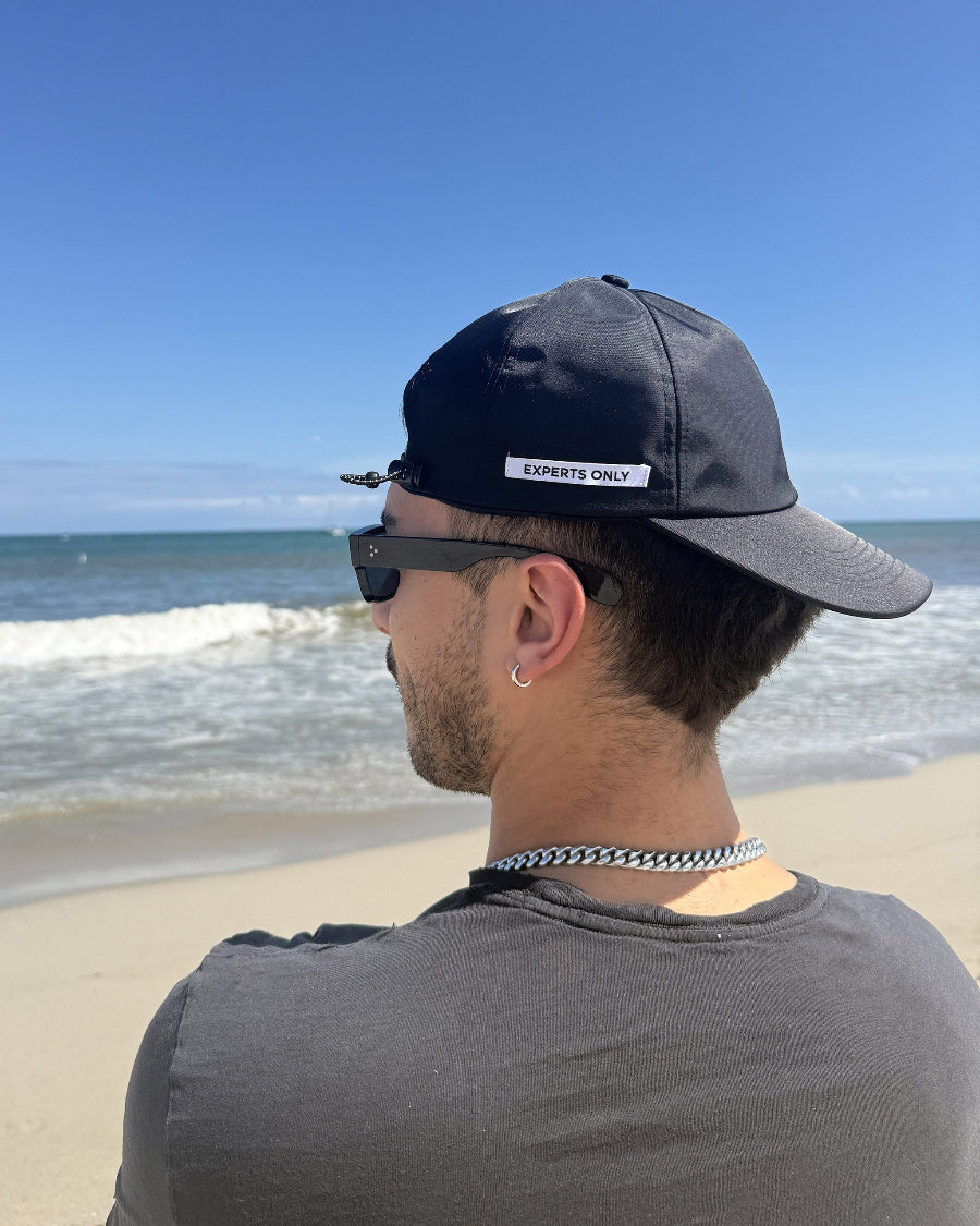 A man wearing a black baseball cap with a white 'EXPERTS ONLY' label, sunglasses, and a silver chain necklace, standing on a sandy beach with the ocean and blue sky in the background.